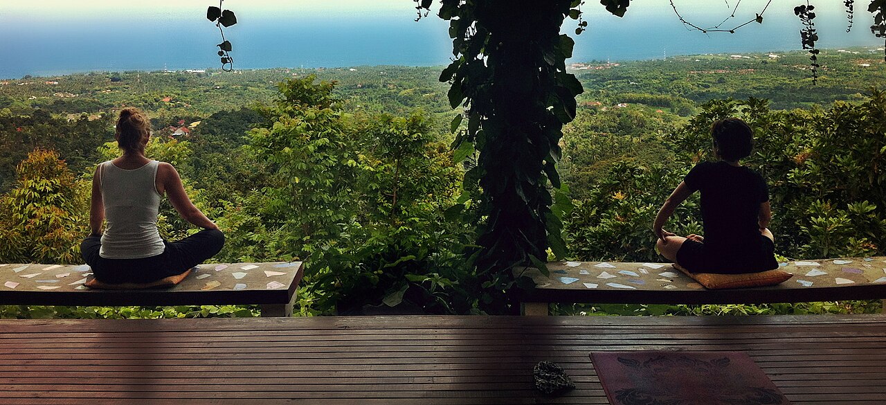 Zwei Personen sitzen im Schneidersitz auf einer Terrasse mit Blick über Wald und Küste Balis