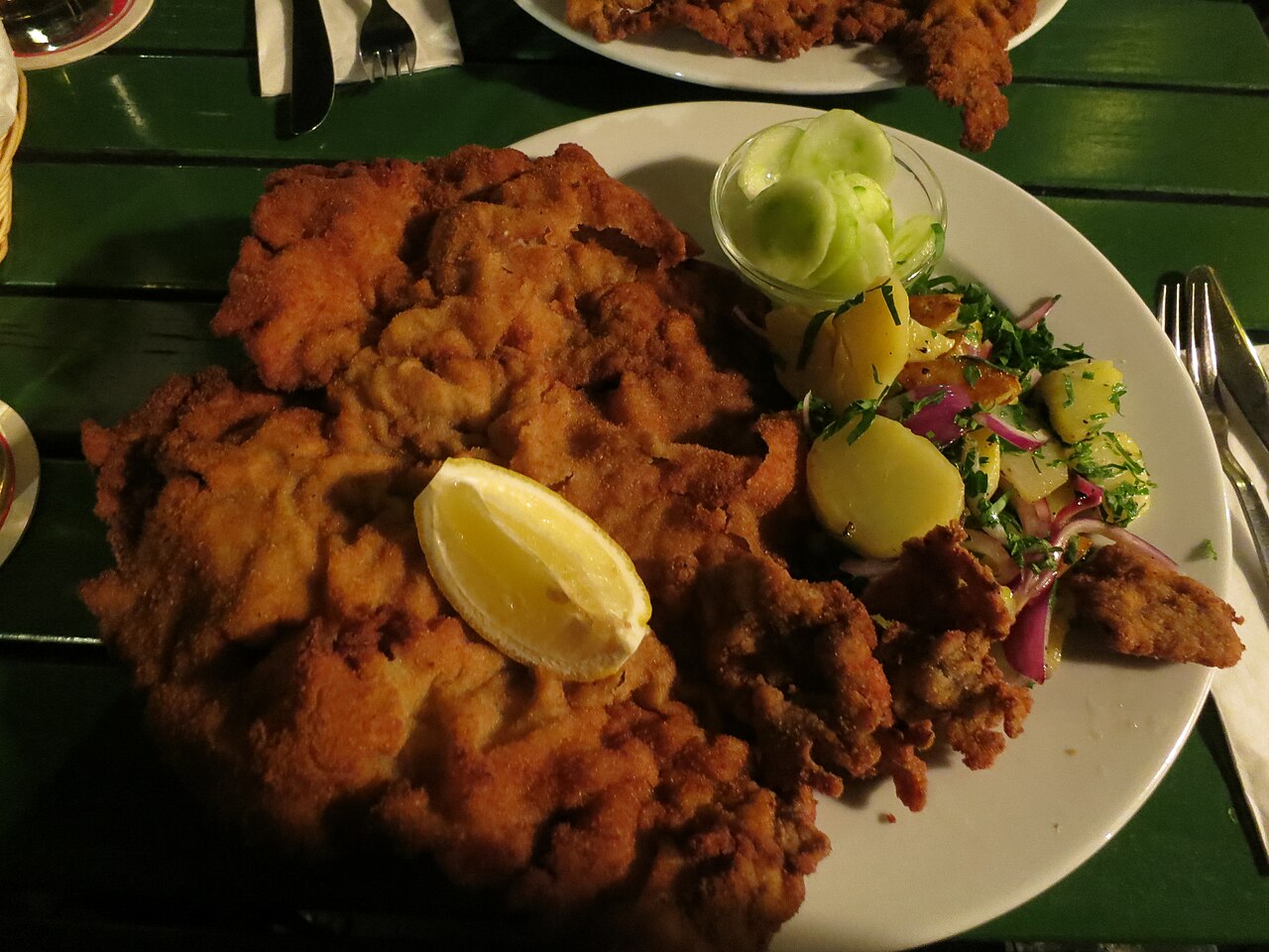 Großes paniertes Schnitzel mit Zitronenspalte, daneben Kartoffelsalat und eine Schale Gurkensalat