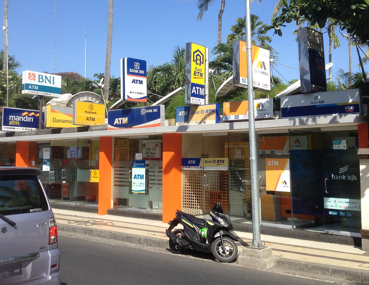 Reihe von Geldautomaten-Kabinen verschiedener Banken an einer Straße in Kuta, davor ein Motorroller