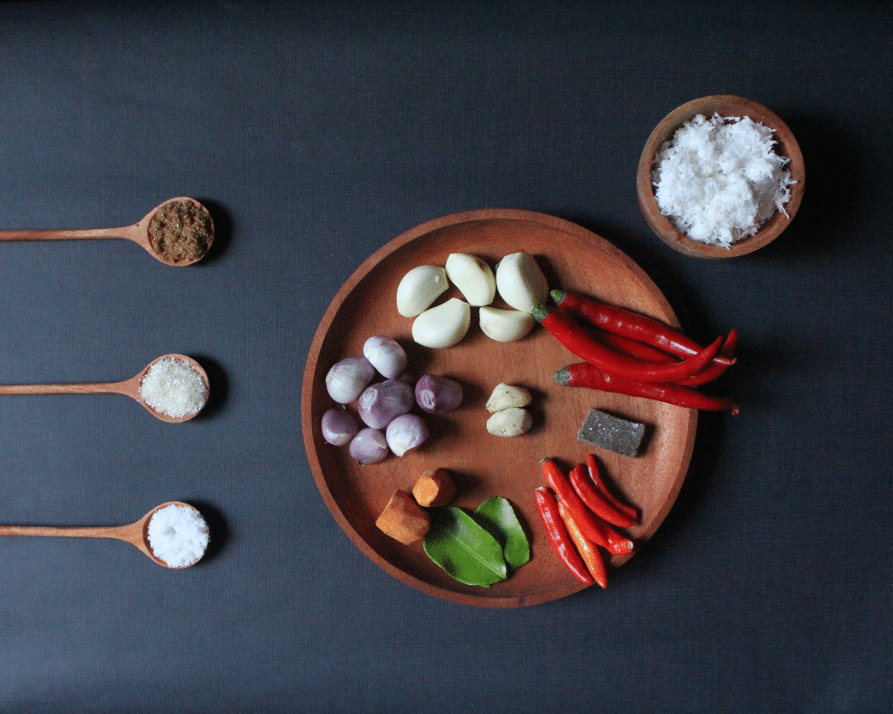 Holzteller mit Knoblauch, Schalotten, roten Chilis, Kencur, Limettenblättern, daneben Kokosraspel und Gewürzlöffel