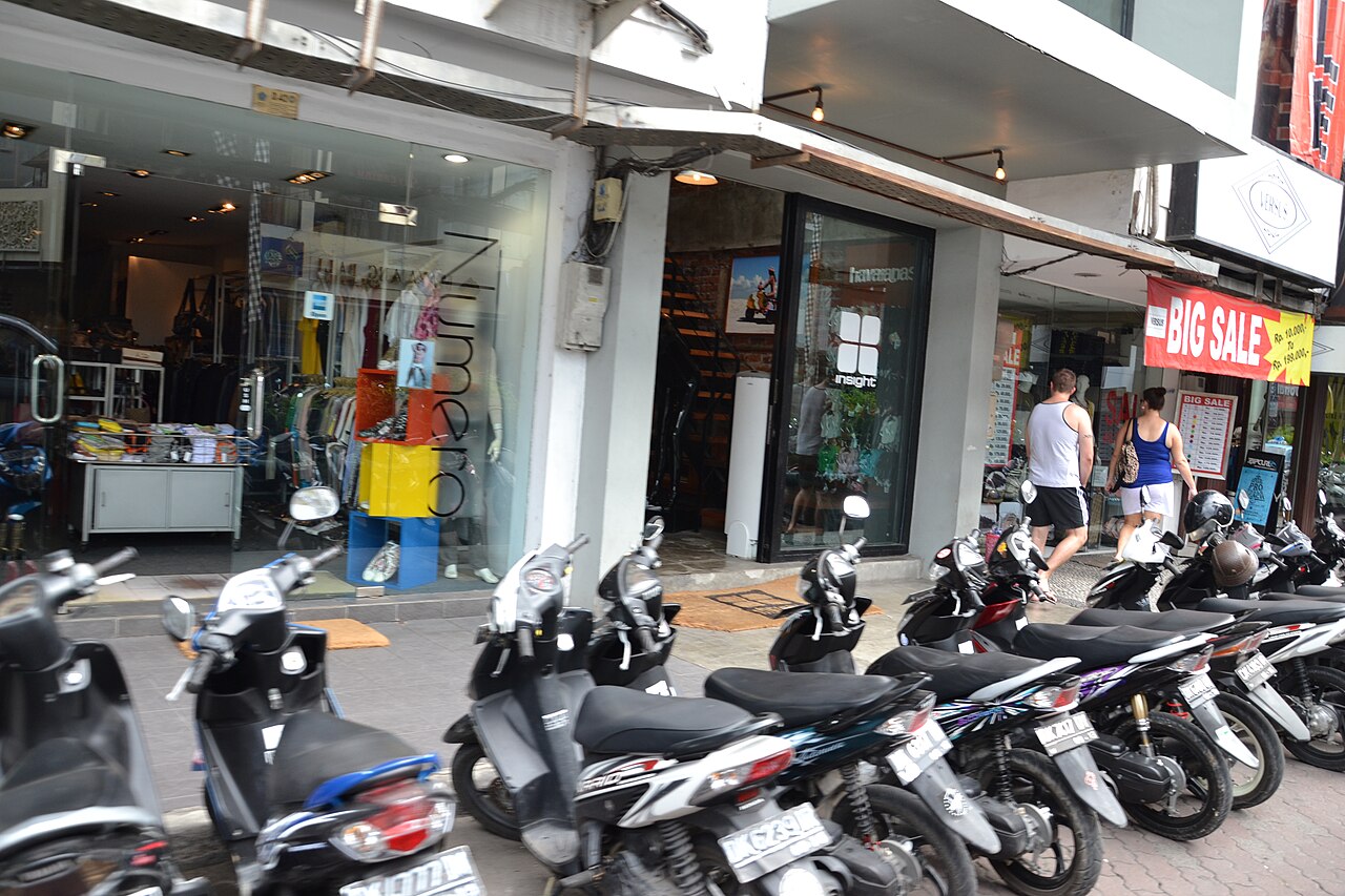 Reihe geparkter Motorroller vor Läden am Kuta Square auf Bali, Passanten auf dem Gehweg