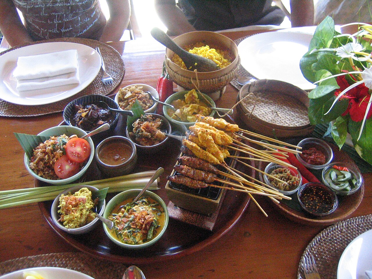 Holztablett mit Sate-Spießen auf Mini-Grill, gelbem Reis, Sambal und Schalen mit balinesischen Beilagen