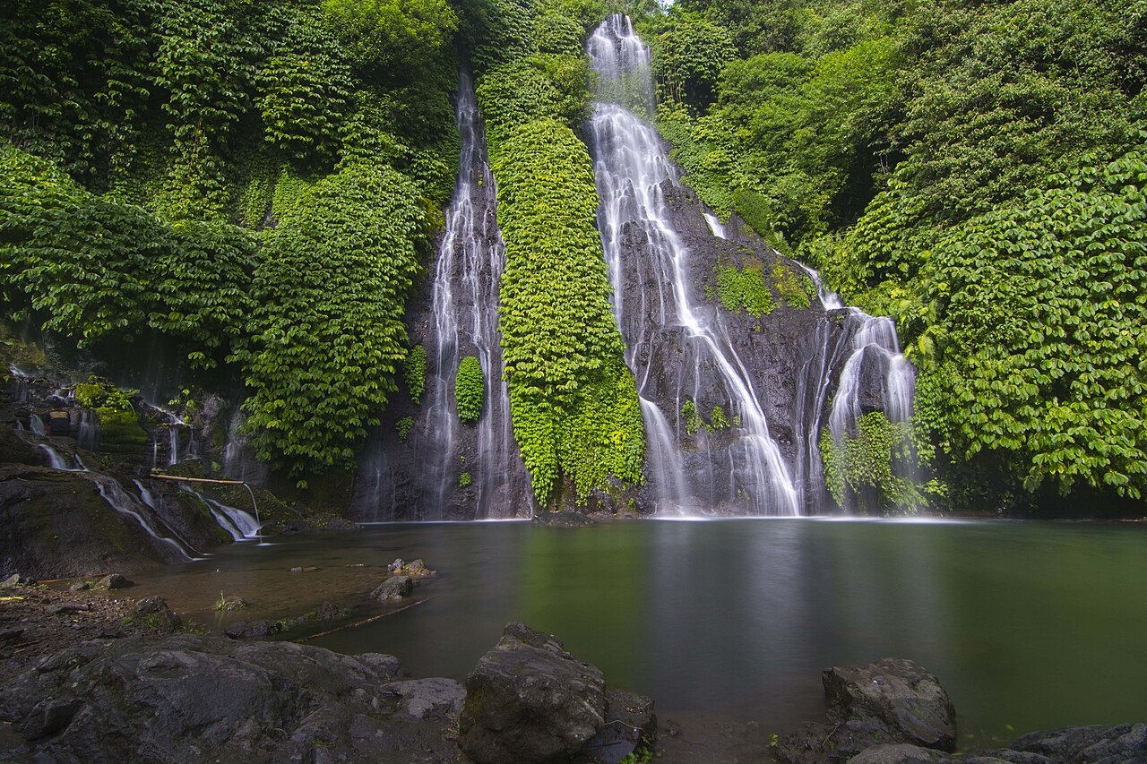 Mehrere Wasserfälle der Banyumala-Fälle an einer grün bewachsenen Felswand über einem ruhigen Becken