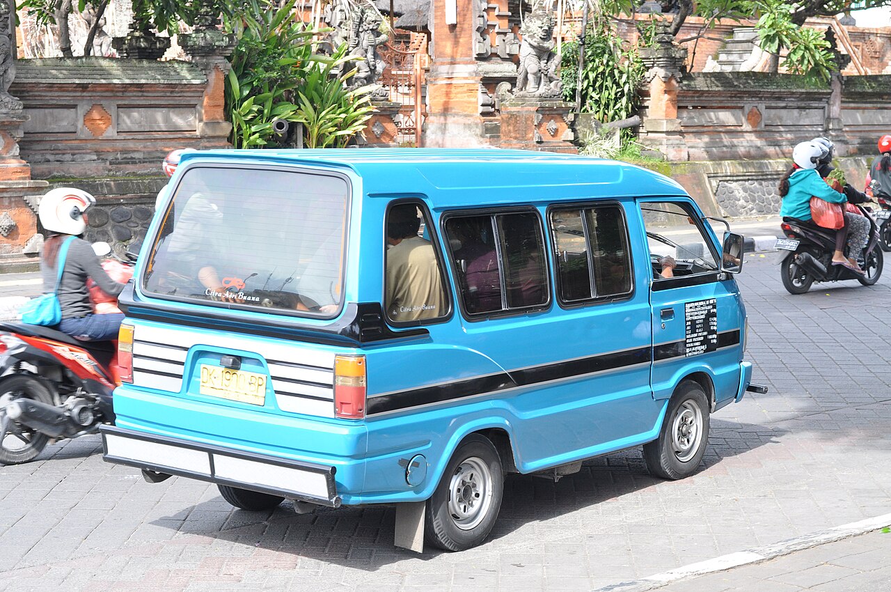 Hellblauer Kleinbus (Bemo) als Sammeltaxi auf einer Straße in Denpasar, daneben Motorroller