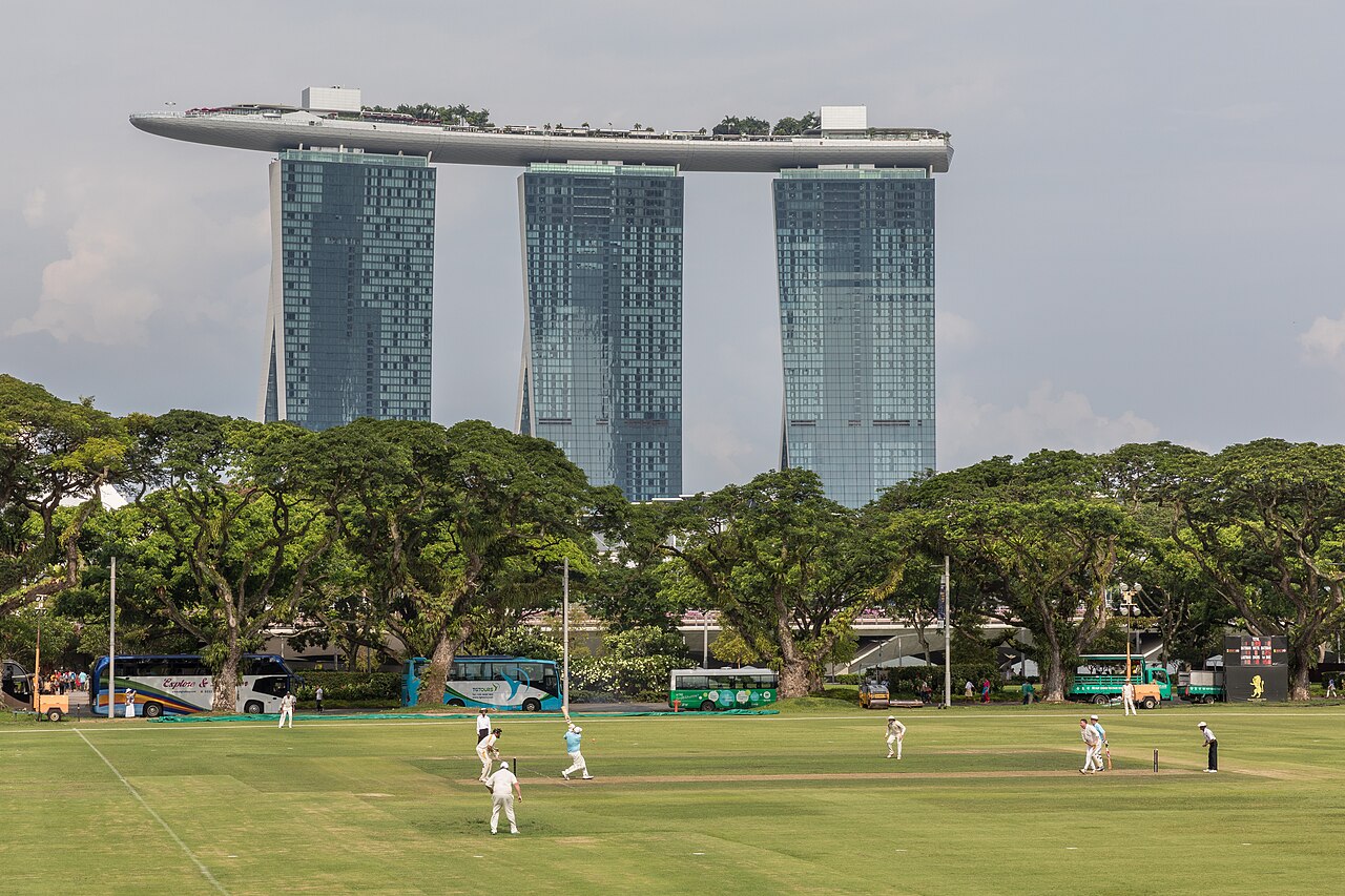 Cricketspieler auf der Rasenfläche des Padang vor Bäumen, dahinter das Hotel Marina Bay Sands mit Dachplattform