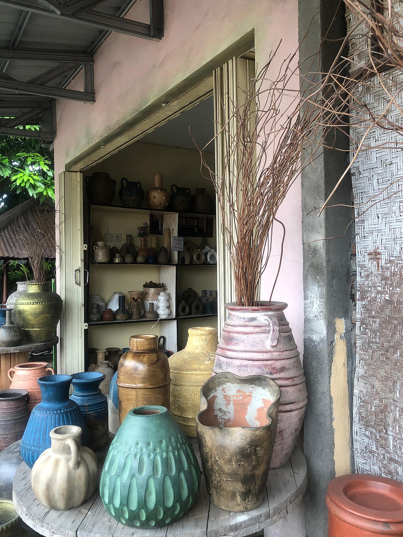 Krüge und Vasen in Blau, Grün, Ocker und Terrakotta vor einem Laden mit Regalen voller Keramik in Banyumulek