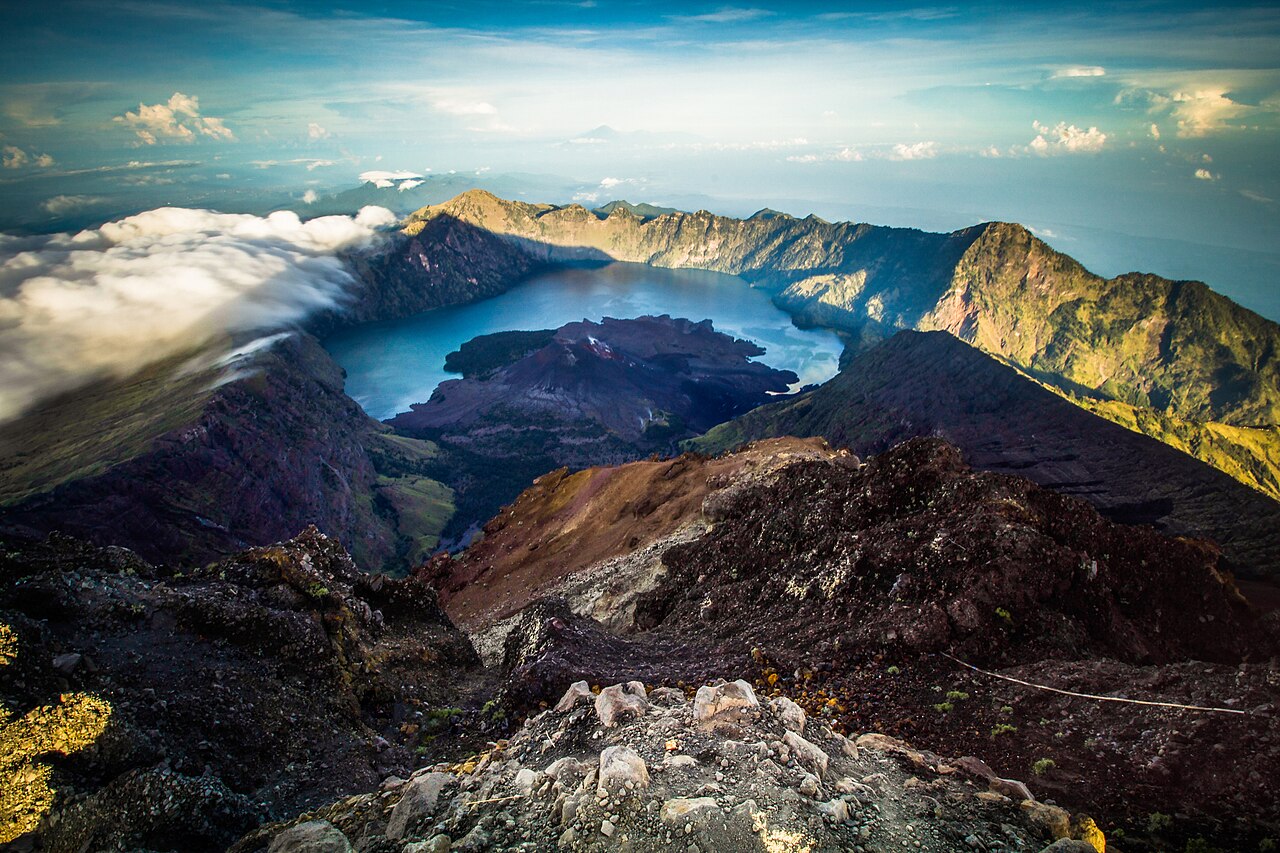 Aussicht vom Rinjani-Gipfel über felsiges Gelände auf die Caldera mit dem Kratersee Segara Anak und Wolken