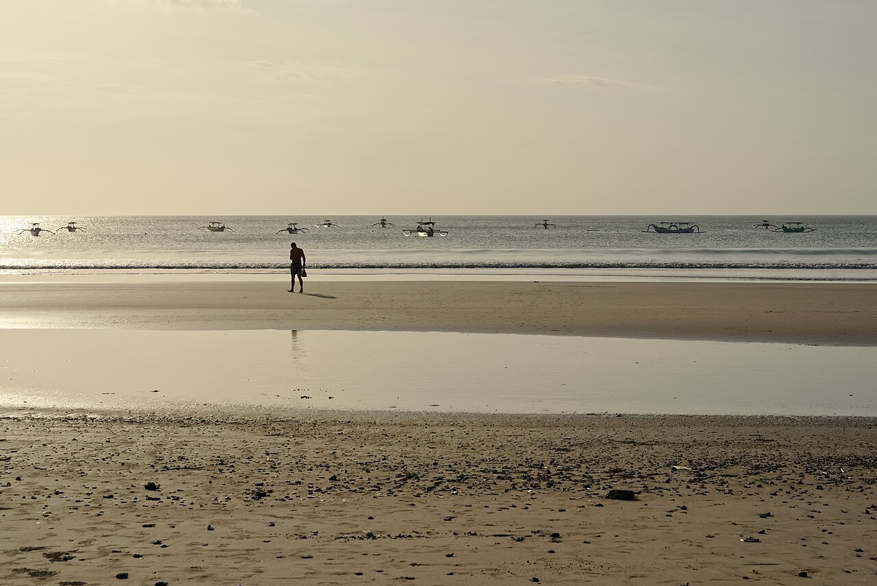 Weiter Sandstrand von Kuta bei Ebbe im Gegenlicht, eine Person am Wasser und Auslegerboote auf dem Meer