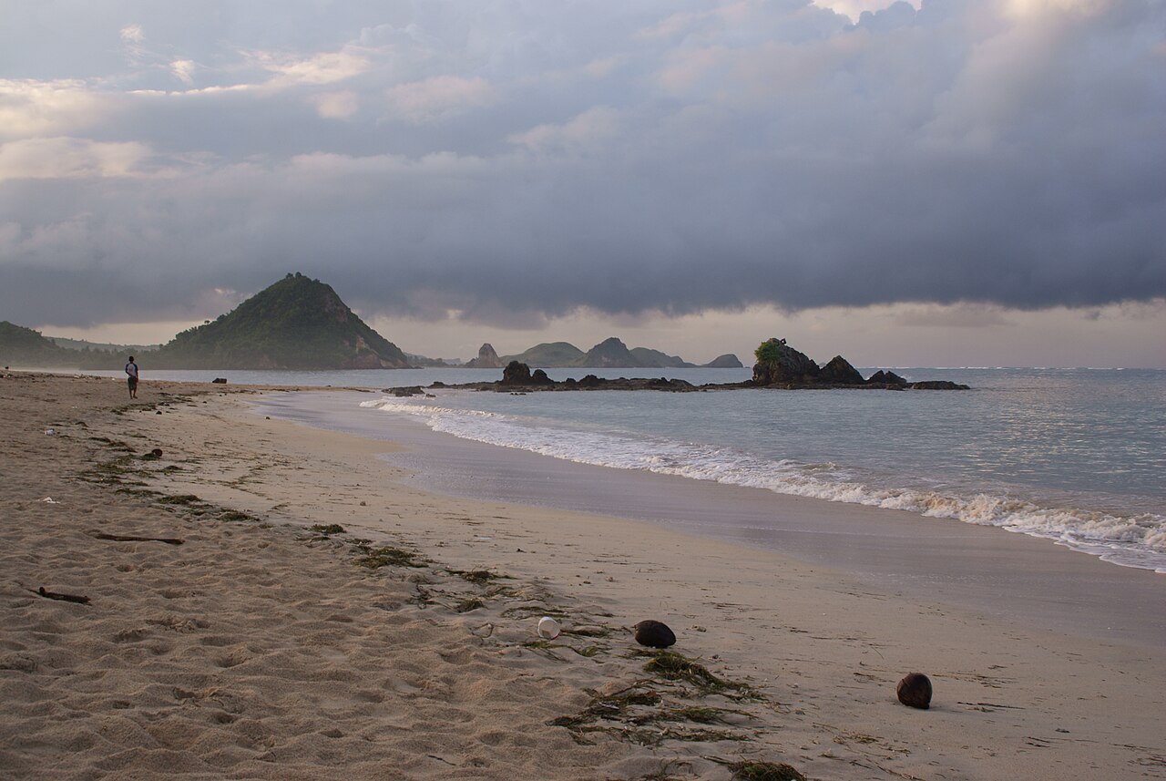 Sandstrand von Kuta Lombok bei bewölktem Himmel, im Hintergrund ein Hügel und kleine Felsinseln