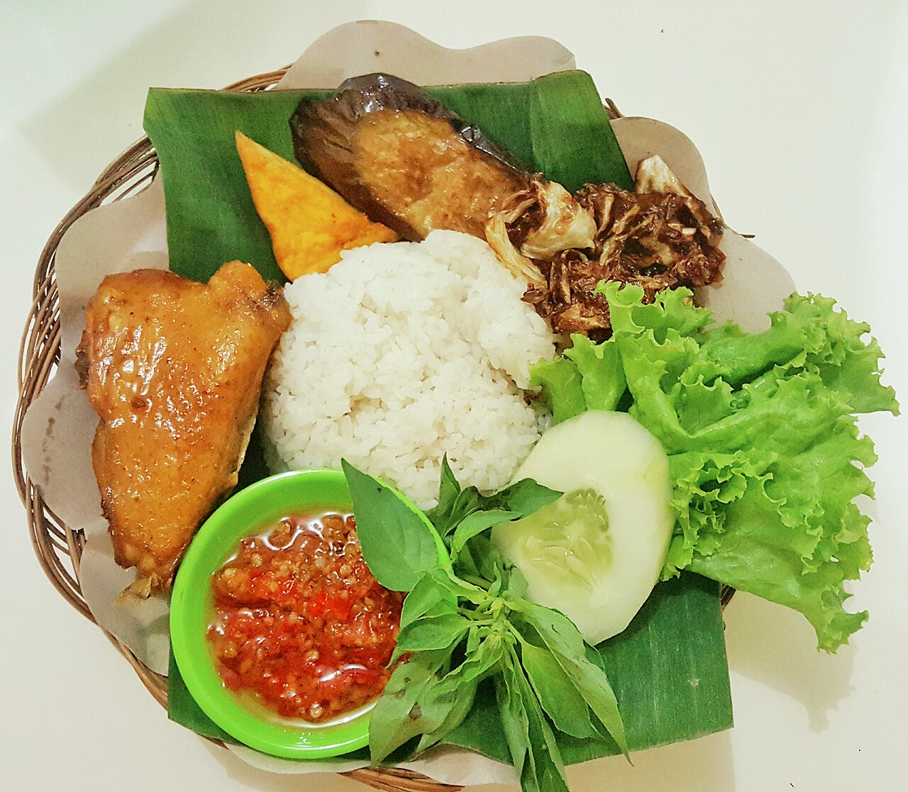 Korb mit Bananenblatt, Reis, gebratenem Hähnchen, Salat, Gurke, Basilikum und Schale mit rotem Sambal