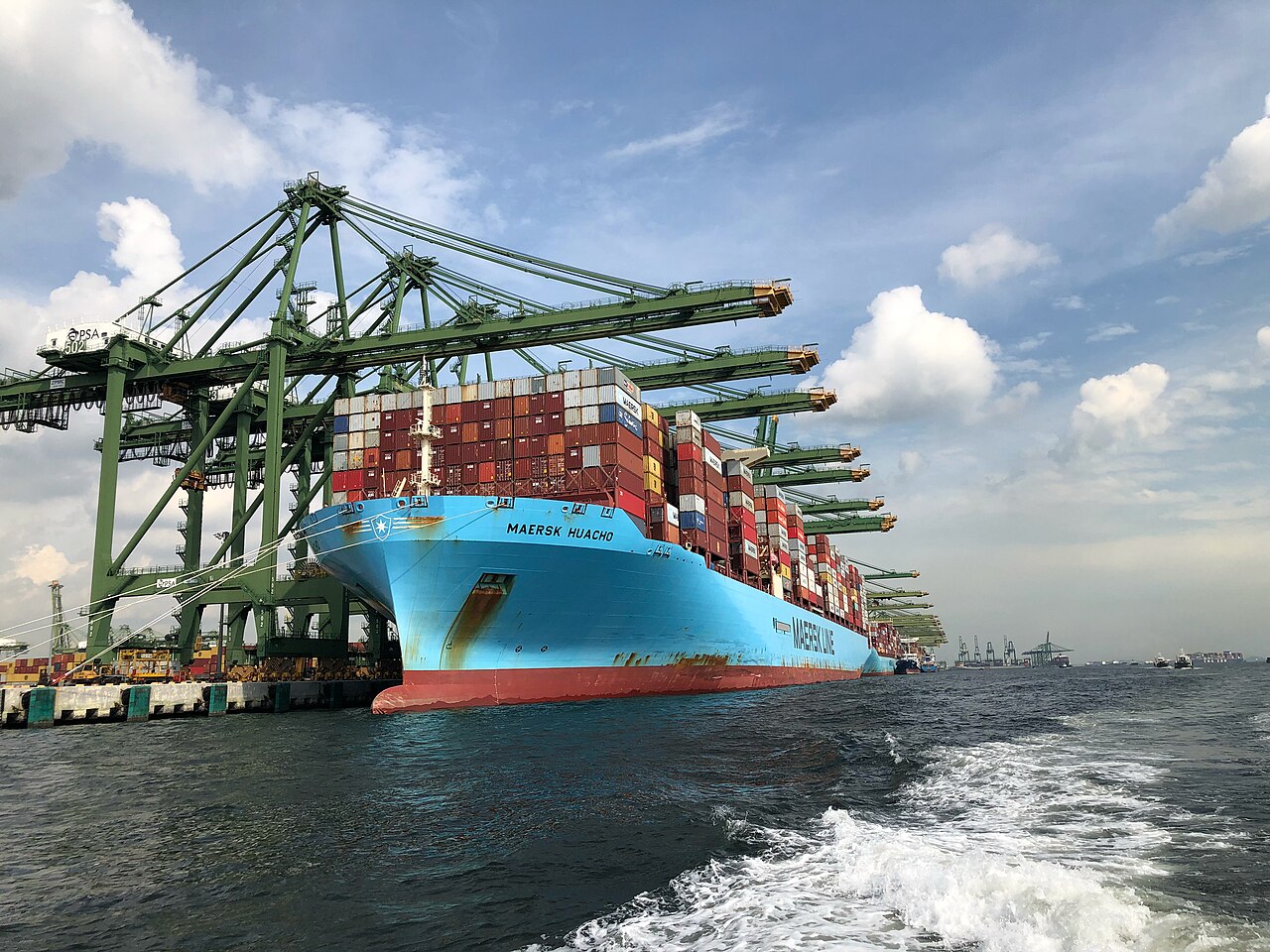 Hellblaues Containerschiff mit gestapelten Containern unter grünen Containerbrücken am Terminal Pasir Panjang