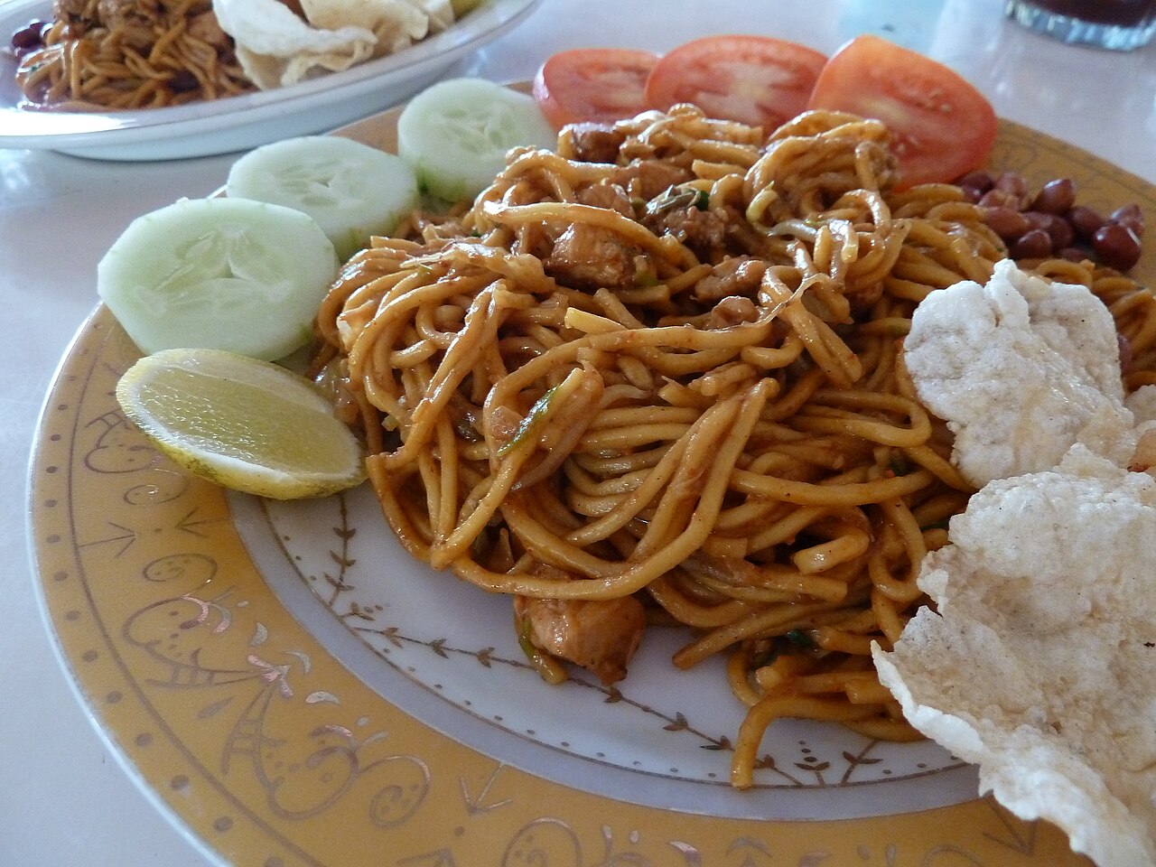 Teller Mie Aceh mit dicken gewürzten Nudeln, Gurkenscheiben, Tomaten, Limette und Krupuk