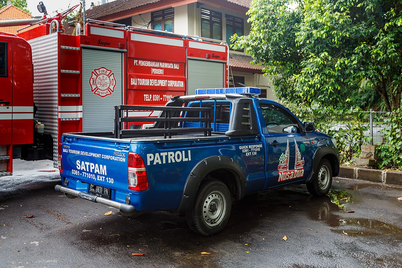 Blauer Pick-up mit Aufschrift Satpam Patroli und Blaulicht, dahinter ein rotes Feuerwehrfahrzeug in Nusa Dua