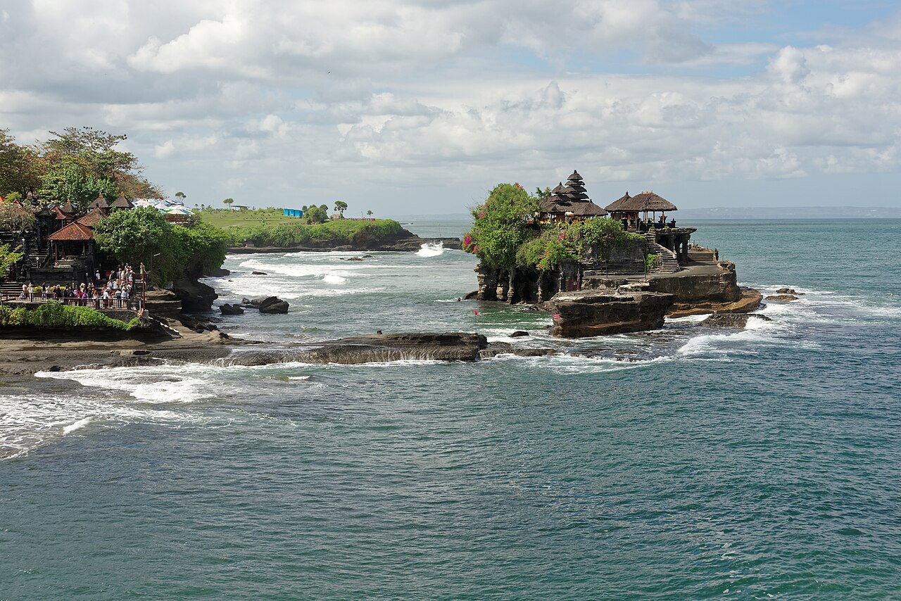 Pura Tanah Lot auf einem Felsen in der Brandung, links Besucher an der Küste, blauer Himmel mit Wolken
