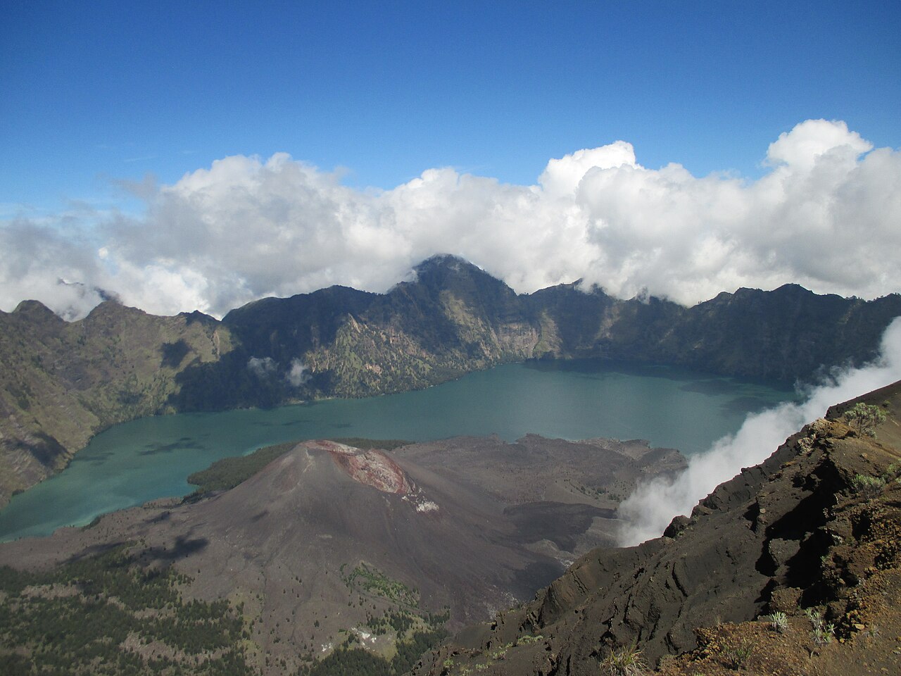 Türkisfarbener Kratersee mit dunklem Vulkankegel davor, umgeben von steilen Kraterwänden und Wolken