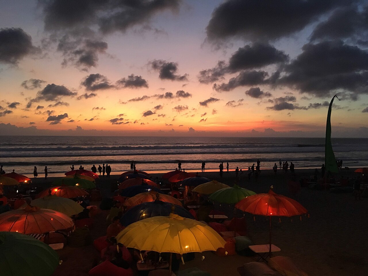 Bunte beleuchtete Sonnenschirme und Sitzsäcke am Strand, dahinter Menschen am Wasser und Abendhimmel mit Wolken