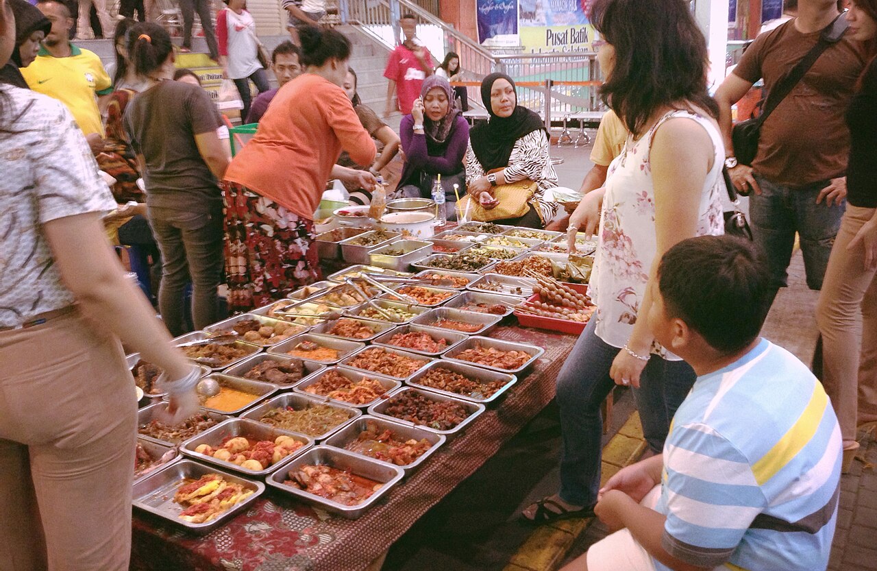 Zahlreiche Schalen mit indonesischen Gerichten auf einem Tisch, Gäste stehen und sitzen um den Stand herum