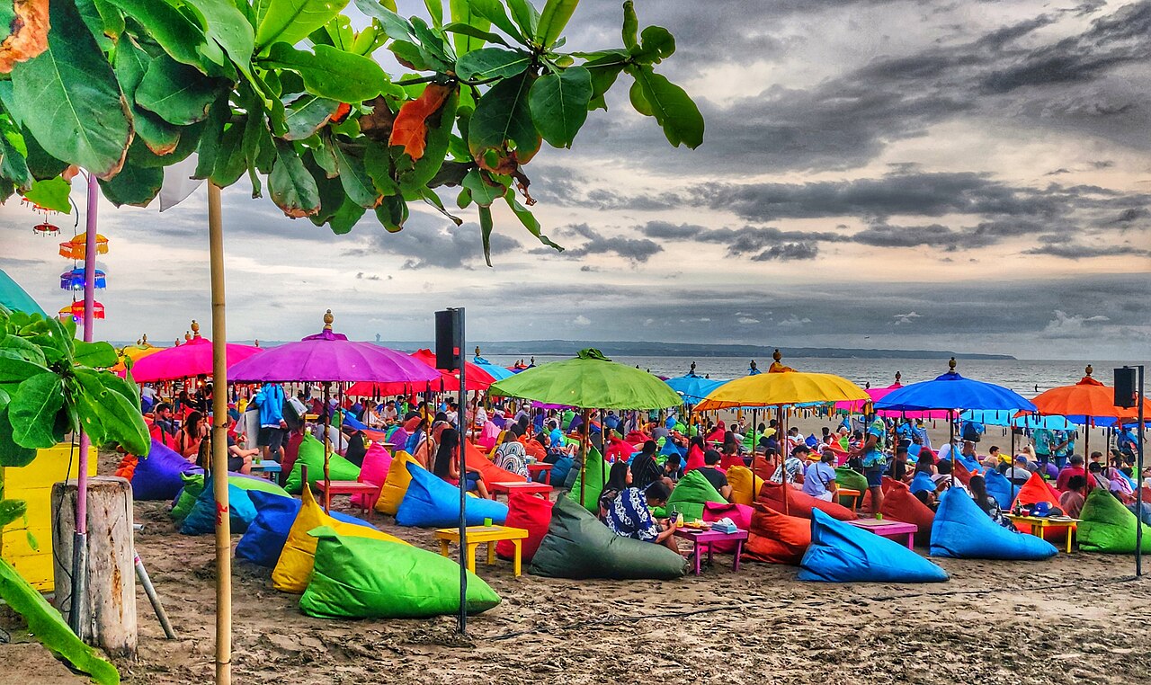 Gäste auf bunten Sitzsäcken unter farbigen Sonnenschirmen an einem Strand auf Bali, Himmel bewölkt