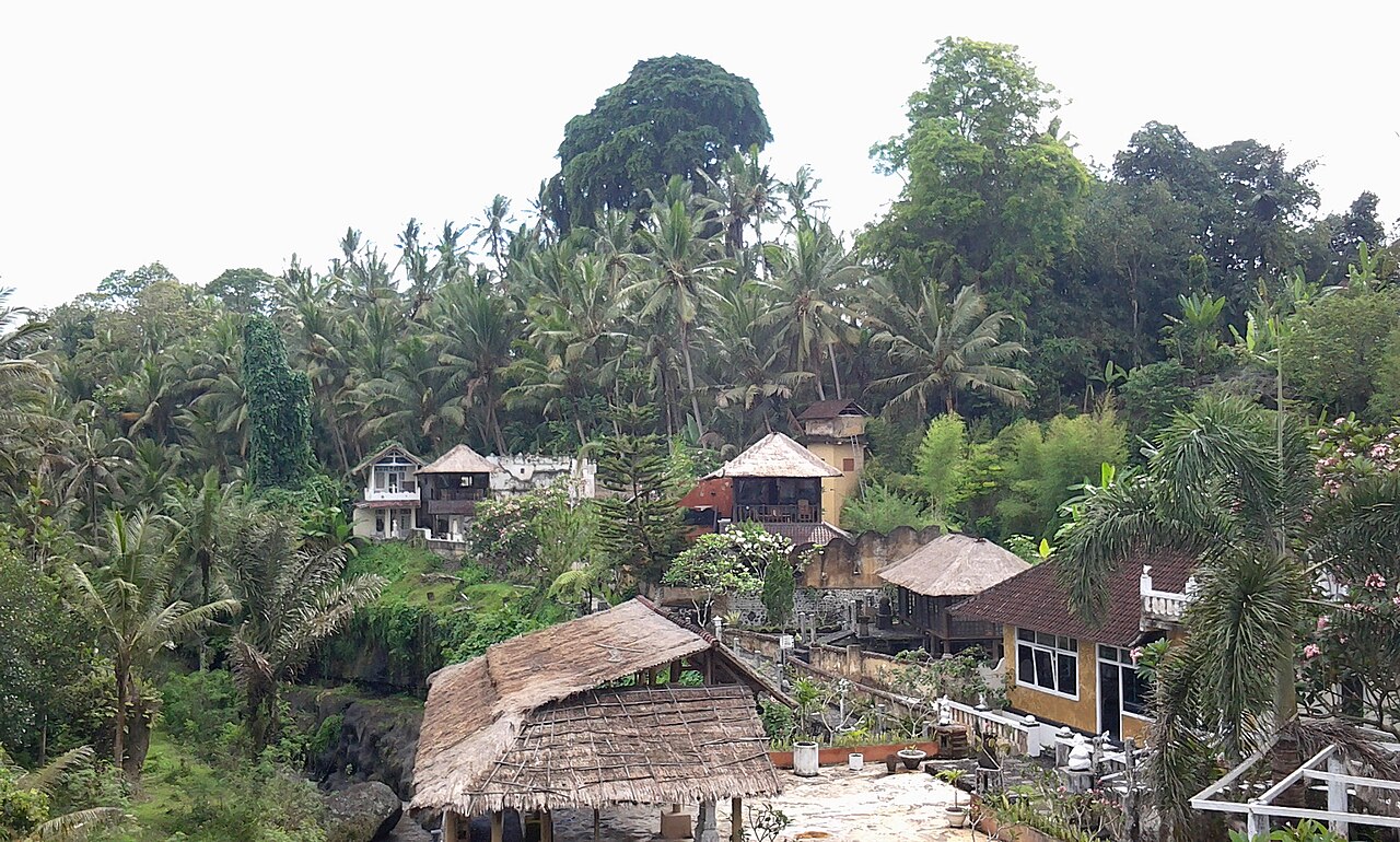 Mehrere Wohnhäuser und Villen mit Ziegel- und Strohdächern zwischen Palmen an einem grünen Hang auf Bali