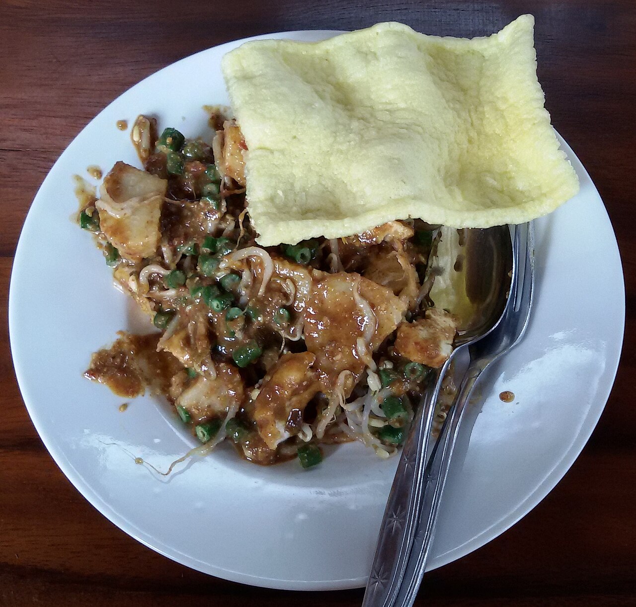 Teller mit Ketupat-Stücken, Bohnen und Sprossen in Erdnusssauce, obenauf ein gelber Krupuk